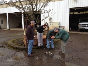 Four people are outdoors, in a muddy area, looking at something on the ground near a planter.