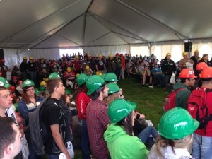 A large group of people, mostly young, wearing hard hats, are seated and standing in a tent.